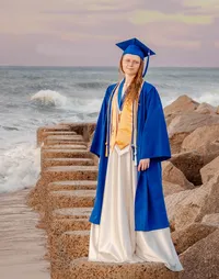 A college senior in a blue cap and gown standing on beach rocks with waves behind in Wilmington, North Carolina. This striking photo highlights the celebration of graduation, perfect for capturing memorable senior moments by the sea.