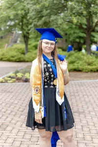 A senior proudly wearing a blue cap and gown with honor cords, standing on a paved path at Cape Fear Community College in Wilmington, North Carolina. This image highlights the achievements of graduation, perfect for students looking for artistic senior photography options.