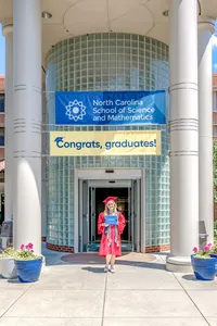 A senior in a red gown holding a diploma in front of the North Carolina School of Science and Mathematics with a congratulatory banner. This celebratory photo captures graduation success, ideal for capturing meaningful senior photography.
