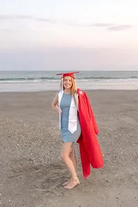 A college senior in a red cap and gown standing barefoot on the beach with the ocean in the background in Oak Island, North Carolina. This joyful image captures the celebration of graduation, perfect for capturing memorable senior moments by the sea.