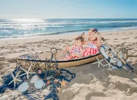Two young children sit in a decorative boat on the sandy beach, surrounded by seashells and nautical props. Dressed in matching red and white outfits, they enjoy a sunny day by the ocean. This image highlights artistic family photography in Wilmington, NC, capturing whimsical childhood moments.