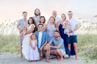 A large, cheerful family gathers on the sandy shores of Carolina Beach, dressed in pastel colors. The group includes parents, children, and grandchildren, all smiling warmly amidst the beach grass and soft evening light. Perfect for those looking for professional family photography in NC.