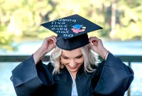 A CFCC graduating senior in a cap and gown, smiling while adjusting her decorated cap at a scenic location in Wilmington, NC. This photo highlights the joy of graduation, perfect for those seeking senior photographer services in Wilmington, NC.