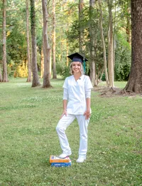 A UNCW senior in a cap and white nursing scrubs standing in a wooded area in Wilmington, North Carolina, with textbooks at her feet. This image captures a blend of academic and career aspirations, perfect for capturing meaningful senior photos.