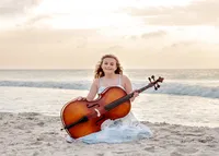 A high school senior in a blue and white dress sitting on the beach with a cello, ocean waves in the background in North Carolina. This unique image highlights the blend of music and nature, perfect for capturing creative senior photography moments.
