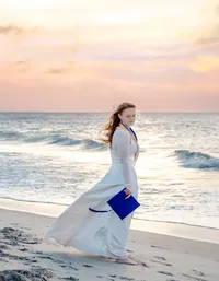 A CFCC senior in a flowing white dress holding her cap with tassle blowing in the wind, walking along Carolina Beach at sunset in North Carolina. This elegant image highlights the celebration of graduation, perfect for capturing memorable senior photos by the ocean.