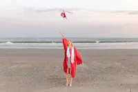 A college senior in a red graduation gown joyfully tossing her cap into the air on the beach with ocean waves in the background in Oak Island, North Carolina. This energetic image captures the celebration of graduation, ideal for vibrant senior moments by the sea.