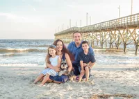 A family sits with their dog on the sandy beach near a wooden pier, dressed in coordinated blue and white outfits. The ocean waves and pier create a picturesque setting. Ideal for affordable family photography in Wilmington, NC, highlighting cherished family moments.
