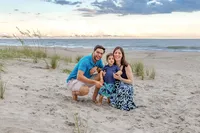 A small family kneels on the beach at dusk, surrounded by sea grass and a calm ocean. Dressed in blue attire, they share a joyful moment, perfect for artistic family photography in Wilmington, NC, highlighting peaceful family interactions.