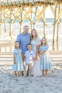 A family of five stands under a pier, dressed in blue and white outfits. The ocean waves and sandy beach provide a picturesque backdrop. Ideal for destination family photographer NC, showcasing elegant and serene beachside portraits.