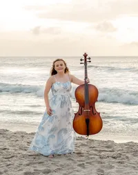 A high school senior in a patterned dress standing with a cello on the beach as waves roll in Carolina Beach, North Carolina. This creative portrait captures the harmony of music and the ocean, ideal for capturing memorable senior photos.