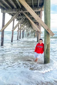A high school senior wearing a red hoodie standing under a pier with waves crashing around in North Topsail Beach, North Carolina. This dynamic photo highlights the adventurous spirit of senior photography, ideal for capturing unique beach moments.