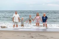 A family of five holds hands and plays in the surf at Carolina Beach, dressed in casual summer attire. The playful interaction with the waves highlights joyful family connections. Perfect for affordable family photography in Wilmington, NC.