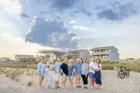 A family stands on the beach in front of beachfront homes, under a dramatic cloud formation. Dressed in blue and white, they enjoy the coastal scenery. Perfect for professional family photography in NC, highlighting beachside gatherings and stunning skies.