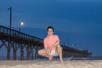 A high school senior in a pink shirt and shorts crouching on the beach at night with a pier illuminated behind in North Carolina. This atmospheric portrait captures the unique charm of senior photography, perfect for evening beach sessions.