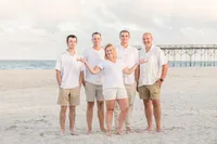 A family of five stands barefoot on the beach near a pier, dressed in coordinated white and khaki outfits. The open sky and gentle ocean waves create a serene backdrop. Ideal for North Carolina family photography, capturing classic beachside portraits.