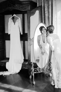 Black and white photo of a bride getting ready by an antique mirror, her gown hanging nearby