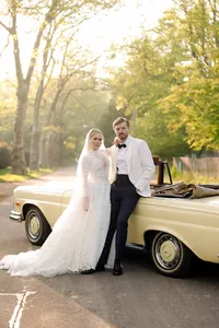 Groom in a white dinner jacket and bride in lace beside a vintage car at golden hour