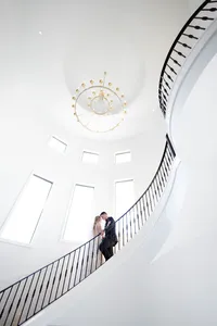 Bride and groom on a sweeping spiral staircase in a white rotunda beneath a chandelier