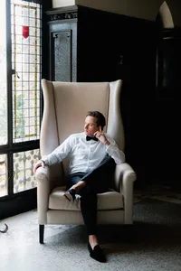 A refined composition of a groom in a tuxedo seated in a high-back leather armchair, reflecting an intentional presence and editorial style.
