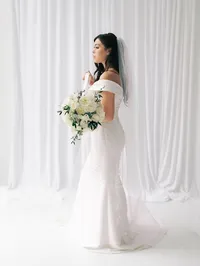 Bride holding a white rose bouquet with greenery in front of a white draped backdrop