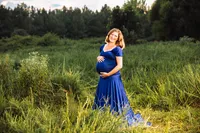 A redheaded pregnant woman in a royal blue dress stands in a large overgrown field on a cloudy day. The landscape is vast.