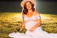 A pregnant woman in a cream fedora sits on short cut grass. It is dusk and she is backlit with small fireflies glistening around her.