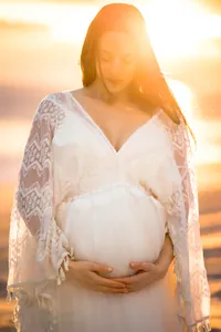 The sun provides a golden backlight to a pregnant mother on the beach. She wears a white dress and looks softly down at her bump while holding it from beneath with both hands.
