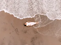 An aerial photograph of a dark haired pregnant woman laying on the beach. The water is cusping the shore and wrapping around her feet.