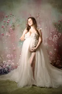 A pregnant woman in a tulle dress smiles up and out of the frame. She has one arm bent against her chest with her hand in her hair while the other arm drapes around her bump, holding it from beneath. She stands in front a multi-colored floral backdrop.
