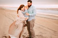 A pregnant mother smiles with her son as they press their foreheads together. Her husband smiles at them while holding he son from behind. The three are on a golden beach shortly after sunrise. The sky and water are pale blue with subtle hues of pink.