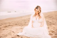 A pregnant woman with blonde hair sits on the beach in a white dress. The shoreline runs diagonally to her and the sky is subtly pink.