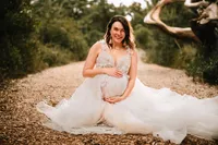 A pregnant woman in a princess style gown smiles with her arms above and below her bump. She sits on a mulched path with bushes on either side. A curved tree shows in the background.