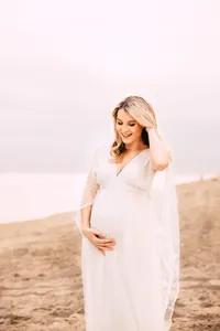 A blonde pregnant woman stands on a beach. She smiles brightly towards her bump. She holds one hand underneath it while fluffing her hair with the other. The sky and beach are bright on an overcast day.