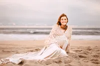 A redheaded pregnant woman sits on a beach. The water and horizon are behind her. The sky is blue with subtle shades of pink.