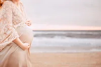 A close picture shows the belly of a pregnant mother peaking into frame with a shoreline and horizon in the background. It is an overcast day, but subtle pink hues can still be seen amongst the gray blue sky.