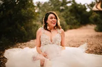 A pregnant woman in a tulle dress sits on a mulched path. She smiles up at someone not seen in the frame.