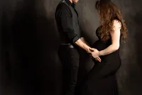 A close up picture of a pregnant woman with her husband on a gray backdrop. She holds his hands to her belly and holds one of her legs slightly forward in the frame. Her long brown hair covers her face, and her husband's face is just out of frame.