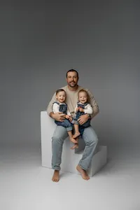 Father sitting on a white studio cube holding his two young children against a soft gray backdrop, timeless studio family portrait captured by Kaitlyn Dawn Photography, professional studio family photographer.