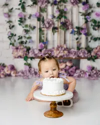Baby leaning forward to taste a white cake during a cake smash session with a purple floral background. Photograph by Yucaipa cake smash photographer Kaitlyn Dawn Photography.
