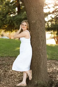 High school senior girl standing beside a tree in a sunlit park wearing a white dress, outdoor senior portrait by Kaitlyn Dawn Photography.