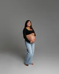 Pregnant woman standing alone in a black top and jeans, smiling while holding her baby bump during a maternity photoshoot in the Inland Empire by Kaitlyn Dawn Photography.