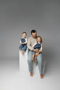 Father sitting on white studio cubes holding his two young children, minimalist gray backdrop Yucaipa studio family portrait by Kaitlyn Dawn Photography.