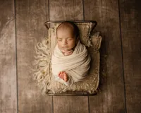 Sleeping newborn baby wrapped in a cream swaddle and posed in a small vintage bed prop on a rustic wooden backdrop during a newborn photography session by Kaitlyn Dawn Photography.