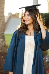High school graduate wearing a blue cap and gown adjusting her cap in warm sunset light, senior graduation portrait by Kaitlyn Dawn Photography.