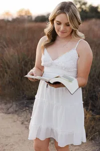 High school senior girl in a white dress reading a book in a golden hour field, outdoor senior portrait by Kaitlyn Dawn Photography.