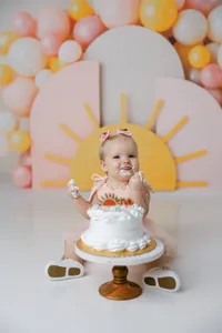 Baby in peach “one” dress enjoying cake smash with sunrise backdrop and pastel balloons. Photograph by Yucaipa cake smash photographer Kaitlyn Dawn Photography.