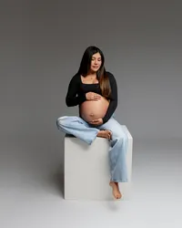 Pregnant mom sitting on a studio block in a black top and jeans, gently holding her belly during a maternity session with Kaitlyn Dawn Photography in Yucaipa.