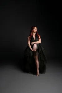 Pregnant woman with long red hair wearing a flowing dark green tulle maternity gown while standing barefoot and gently holding her baby bump during a dramatic studio maternity photoshoot on a dark backdrop captured by Kaitlyn Dawn Photography.