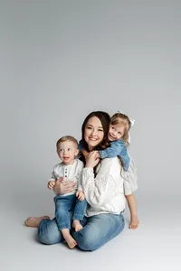 Mother sitting on studio floor holding her two young children close, soft neutral backdrop and natural light creating a warm family portrait by Kaitlyn Dawn Photography, studio family photography specialist.
